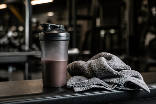 Protein shaker bottle with supplement drink in a dark gym. A grey gym towel rests beside it on a black bench. This image suggests fitness, recovery, and a healthy lifestyle.