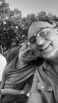 A smiling bald man wearing glasses and a woman pose closely together for a candid selfie outdoors against a backdrop of trees, captured in monochrome.