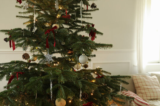 Elegant Traditional Christmas Tree Fir with Red Ribbons, Warm Fairy Lights, Natural Ornaments. Elegant Scandinavian Interior. Festive Holiday Home Decor. Winter Celebration. Blurred window, cushion