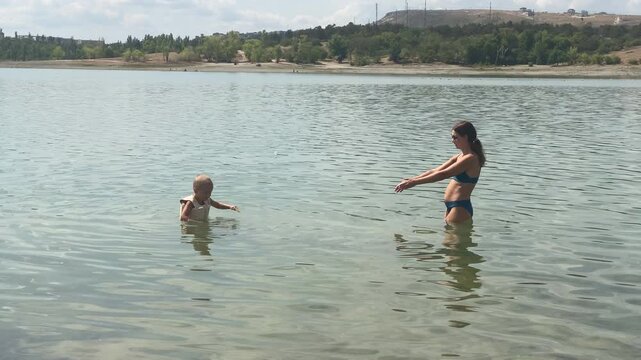 A caring mother reaches out her arms encouragingly toward her young son in the water. Wearing a swim vest, the little boy wades through the clear, calm lake on a warm summer day.