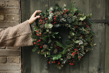 Obraz premium Female Hand in sweater Holding Handmade Christmas Wreath with Evergreen Foliage, Red Berries, Ivy, Eucalyptus on Blurred Old Wooden Door. Festive Winter Holiday Decoration, Natural floral home decor