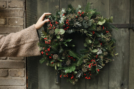 Female Hand in sweater Holding Handmade Christmas Wreath with Evergreen Foliage, Red Berries, Ivy, Eucalyptus on Blurred Old Wooden Door. Festive Winter Holiday Decoration, Natural floral home decor