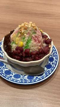 Video top view of Ais Kacang Es Campur shaved ice dessert with red bean, grass jelly, peanuts on wooden table.