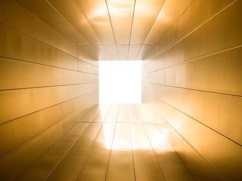 Interior view of a symmetrical tunnel or passage clad in gold-toned metal panels, leading toward a square opening flooded with bright white light. 