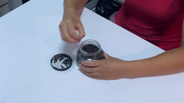 A focused woman carefully places black peppercorns into a jar on a white table. She works calmly in a clean indoor kitchen. The mindful preparation feels peaceful and organized.