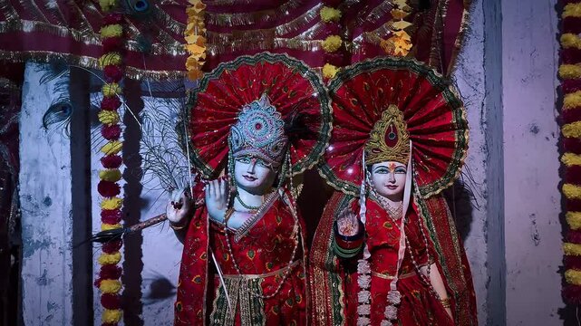 Radha and Krishna displayed side by side, featuring elegant crowns, colorful traditional clothing, flower garlands, and vibrant red ornamental fans that enhance the sacred and festive temple 