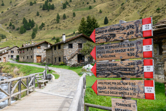 Wooden hiking trail signs beside a footbridge and historic stone cottages in Case di Viso near Ponte di Legno, a scenic gateway to the Italian Alps.