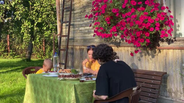 A happy family shares a delicious meal together outdoors. They sit at a green table near a rustic house with vibrant pink flowers. The cozy garden setting feels peaceful and warm.