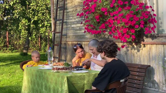 A happy family shares a meal together outdoors. They sit at a green table near a rustic house with vibrant pink flowers. The cozy garden setting feels peaceful and warm.