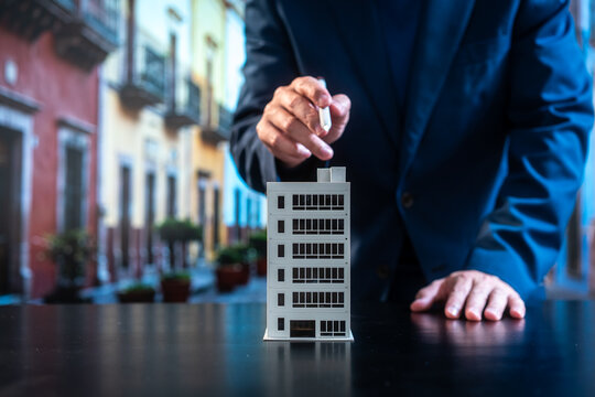 Business professional placing a small office building model on a table, symbolizing real estate investment, urban development, planning and corporate decision-making against a blurred city street.