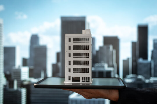 Hand holding a miniature office building on a glass platform against a blurred modern city skyline, symbolizing urban planning, real estate investment, architecture, and business growth.