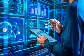Naklejka na ściany i meble IT professional using a tablet and stylus while monitoring glowing blue data dashboards and server systems in a modern technology control room.