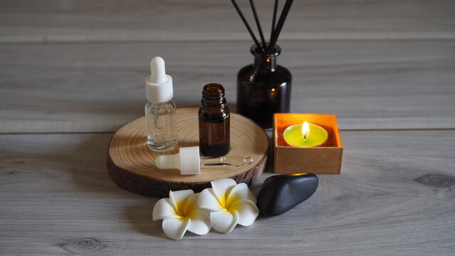 A spa and wellness set, arranged on a natural wood base, includes bottles of essential oils and serums with droppers, a bottle of reed diffuser, a lit scented candle in a wooden box, dark-colored 