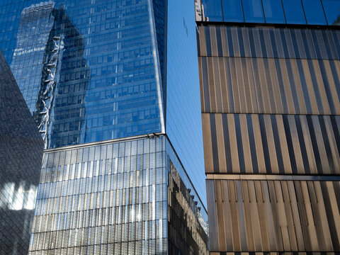 Exterior view of adjacent business office towers in Lower Manhattan, one with a reflective glass facade showing a mirrored building, the other clad in vertical bronze-toned metal fins. 