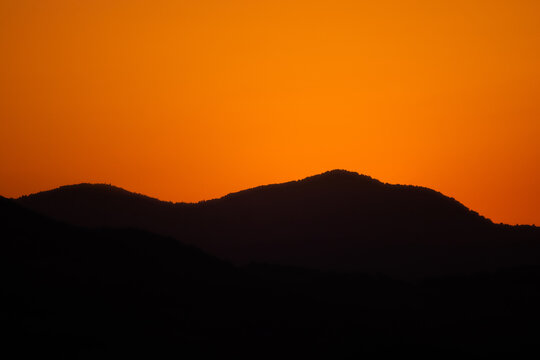 Dark silhouettes of rolling mountain ridges stand out sharply against a brilliant vibrant orange sunset sky, creating a peaceful and dramatic evening mood.