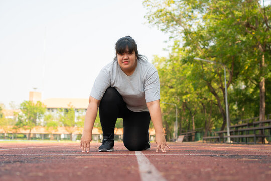 Plus size woman in a sprint start position on an outdoor running track, preparing for exercise with determination and focus in a sunny park, supporting fitness, motivation and healthy lifest