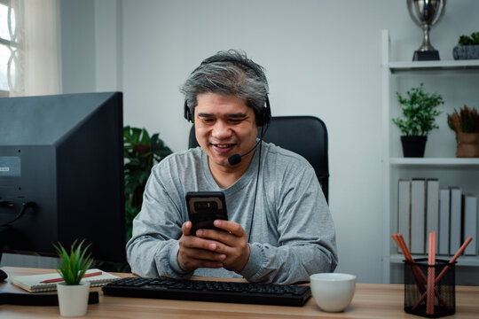 Smiling man wearing a headset and using a smartphone at a computer desk, representing digital communication, remote work, connectivity, and a modern home office lifestyle