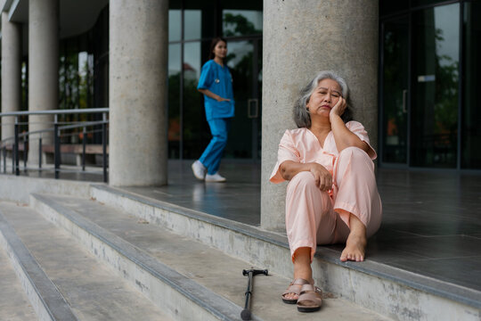 Older woman sits outside a medical facility with a walking cane while a healthcare worker walks in the background, illustrating aging, mobility, healthcare access, and patient support.