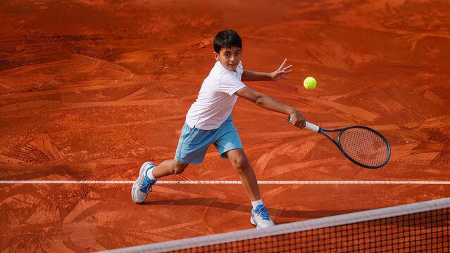 Young Indian boy playing tennis hitting backhand shot on clay court during training match action