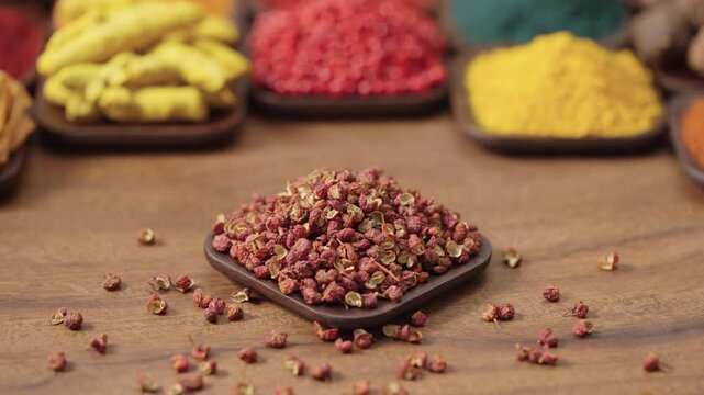 red sichuan peppercorn, open seed pods without seeds in a square bowl on a wooden table, surrounded by various spices and herbs.
