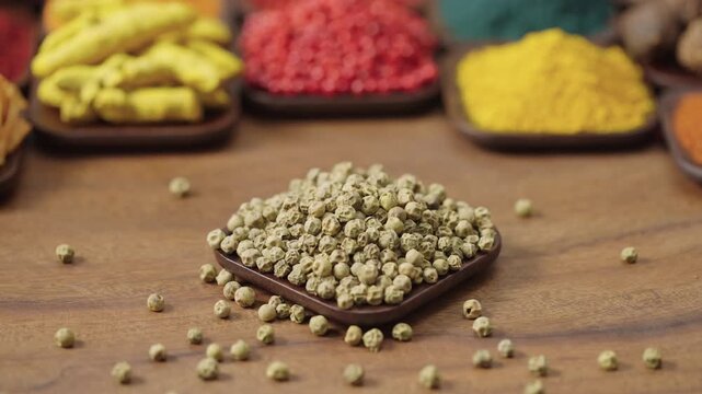 green peas dried unripe pepper plants Piper nigrum in a square bowl on a wooden table, surrounded by various spices and herbs.