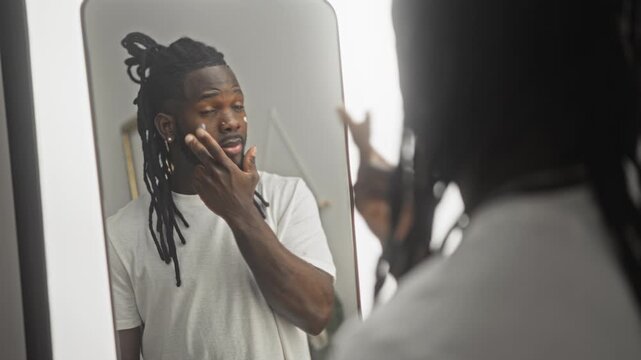 Young african american man applies moisturizer to cheek at illuminated studio mirror while checking skin and hair in white t shirt; self care calm.