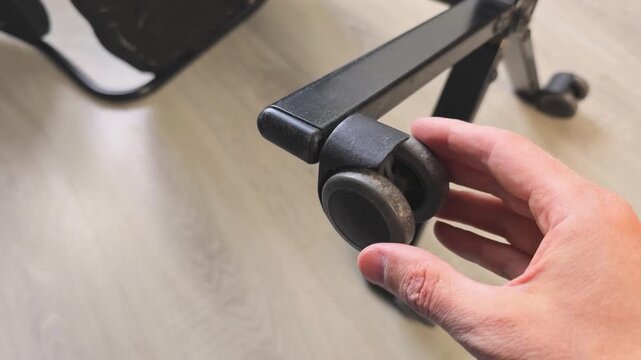 Close-up of a hand inspecting a black plastic-and-rubber caster wheel on an office chair frame; visible dust and buildup on the wheel surface