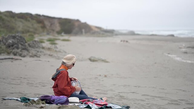 A mature woman plays a drum on the beach while surrounded by sand and natural scenery in Fort Bragg, California.