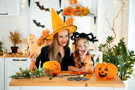 Halloween, a happy family a mother with a baby girl in festive costumes and makeup is preparing for Halloween at home in the kitchen by coloring and carving pumpkins and scaring everyone