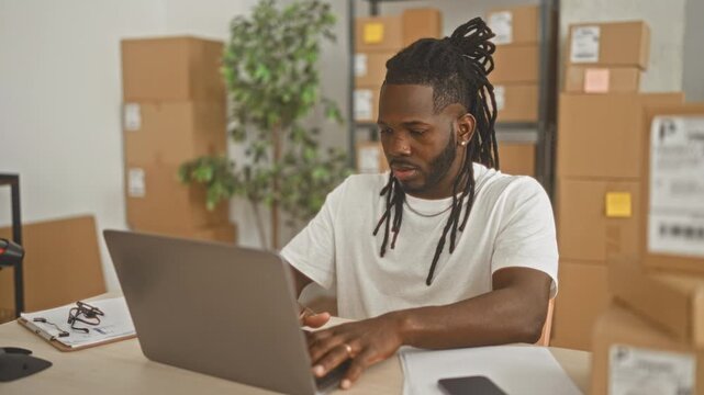 Man typing on laptop at desk surrounded by stacked shipping boxes, clipboard and phone, hands on keyboard in a small packing area inside a building; entrepreneurial focus.