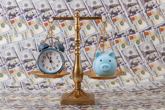 A piggy bank and an alarm clock on the scales against a background of US $100 bills.
