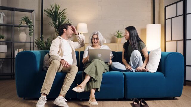 Happy family with a mature mother sitting on the sofa with her son and daughter in law, using a laptop and having a pleasant conversation