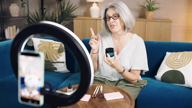 Older woman with grey hair recording a tutorial for her social media channel, reviewing a new beauty cream using her smartphone and a ring light
