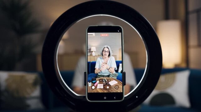 Older female influencer explaining cosmetics products to her audience. Smartphone screen on a tripod with a ring light recording a video