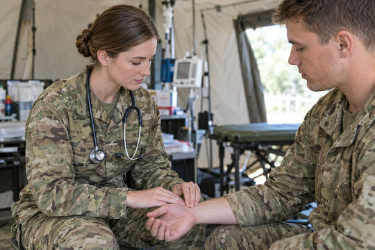 Female military medic in camouflage uniform checking soldier's pulse and hand inside field hospital tent, medical examination scene