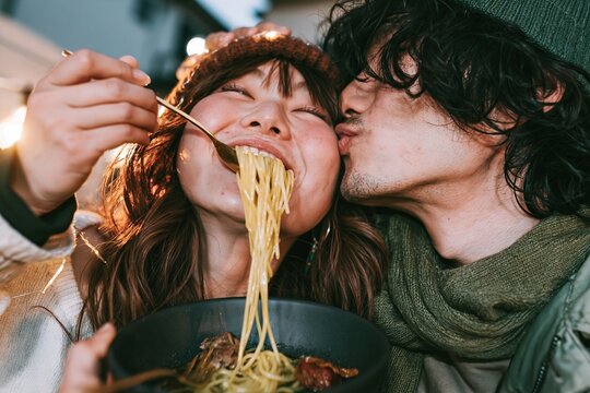 Happy young couple eating noodles together outdoors with the man kissing her cheek affectionately