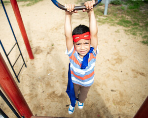 Child in superhero costume hangs from playground equipment outdoors. © Rawpixel.com