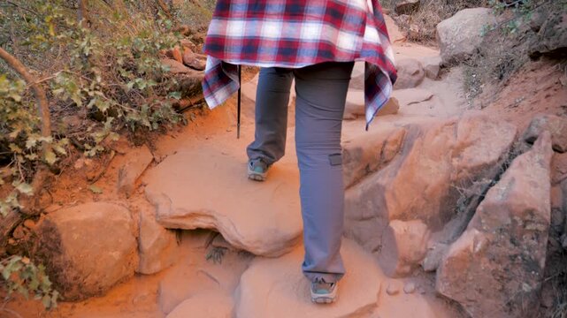 Following woman behind as she carefully hikes rises on stone steep, rocky and sandy trail in canyon. Hiker using trekking poles for stability during outdoor adventure. Rear view legs walking tourist