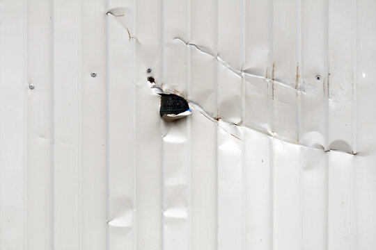 White corrugated metal siding shows denting, cracking, and a puncture hole with dark interior