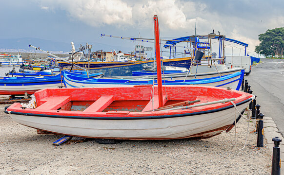A red and white wooden fishing boat rests on the shore near a harbor under a cloudy sky.