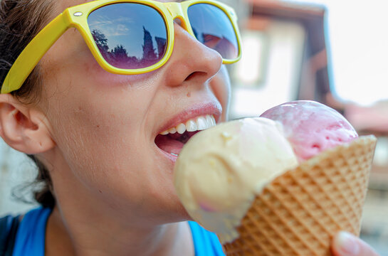 A young woman wearing bright yellow sunglasses smiles as she eats a waffle cone filled with scoops of ice cream outdoors on a sunny day.