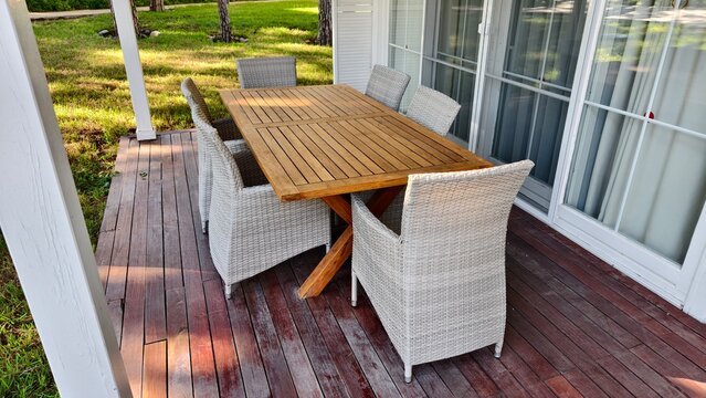 On the wooden terrace with columns there is a wooden table and chairs made of artificial rattan. There are windows and a glass entrance door to the house, a grassy lawn and trees. Sunny autumn weather