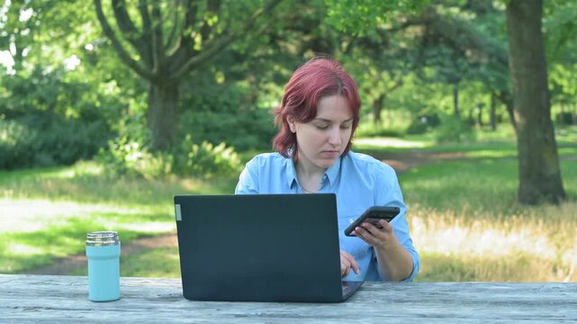 A young ginger woman getting distracted from her laptop work by a smartphone call. She sits at a wooden table in a green summer park, takes her phone, and answers with a smile. Concept of remote busin