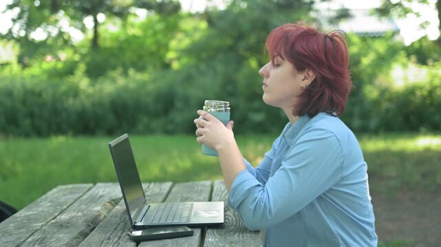 A thoughtful young ginger woman holding a thermal travel mug and drinking hot coffee or tea. She stops working on her laptop at a park table, looking away in deep thought, enjoying a relaxing moment i