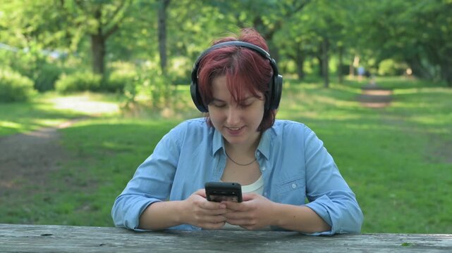 A young woman sitting at a wooden park table, enjoying music in her over-ear headphones while scrolling through social media on her smartphone.