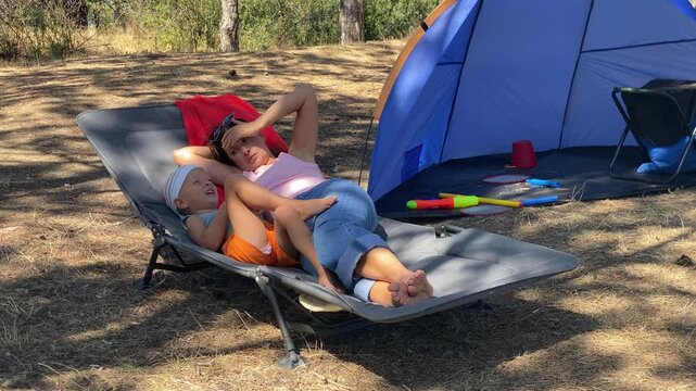 A relaxed mother and cheerful boy lie together on a cozy lounger cot. She rests peacefully while the smiling child cuddles close. They enjoy a warm, serene afternoon at a sunny campsite.