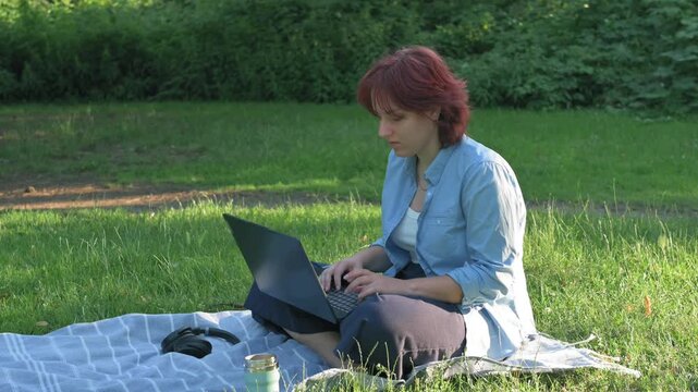 Medium shot of a focused redheaded professional using a portable computer for remote digital marketing or distance education while sitting on a meadow rug during a sunny day.