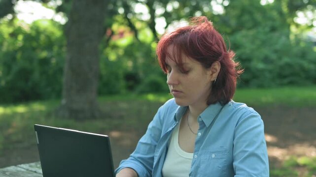 Redhead female typing on her notebook computer, then pausing to pick up her smartphone and dial a number.