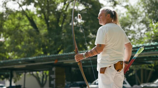Mature man with long hair aiming and shooting an arrow with a traditional wooden bow. Focused athlete practicing archery in a park