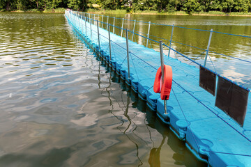 Naklejka premium Long blue floating plastic pontoon bridge with a pedestrian walkway and orange lifebuoy stretching across a calm green lake towards a summer forest.